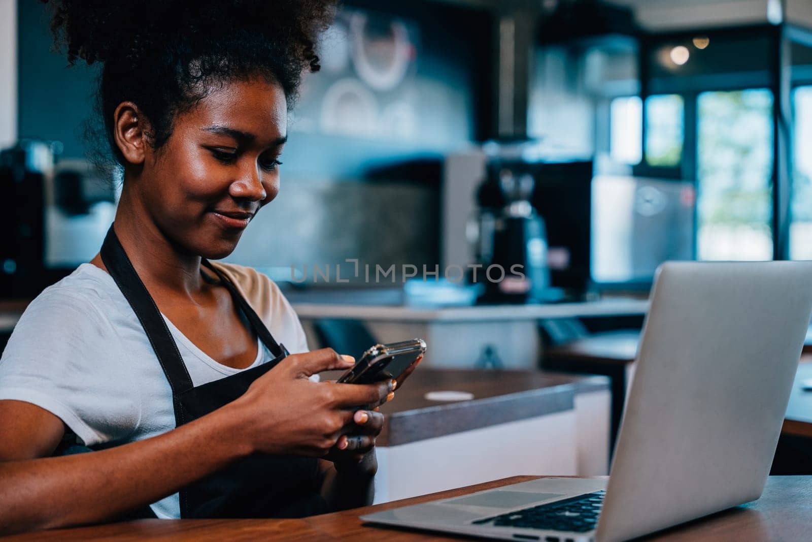 Portrait of cafe owner a concentrated black woman in apron using smartphone in her shop. Multi tasking barista manages orders communicates runs her cafe ensuring excellent service. by Sorapop