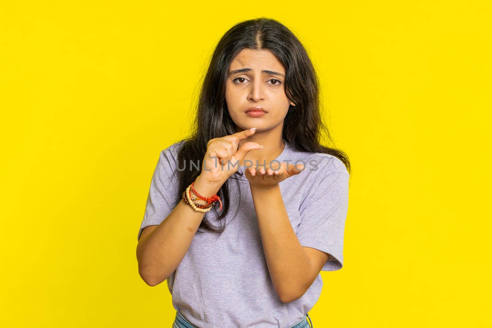 Need some more, please give me. Indian woman showing a little bit gesture with sceptic smile showing minimum sign, measuring small size, begging help. Arabian girl isolated on yellow studio background