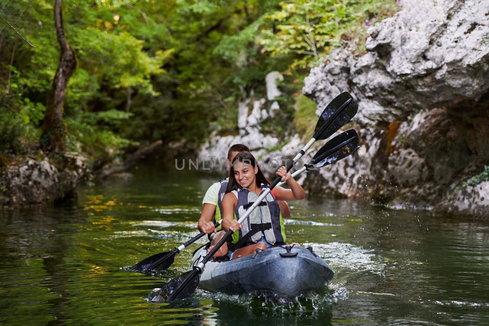 A young couple enjoying an idyllic kayak ride in the middle of a beautiful river surrounded by forest greenery.