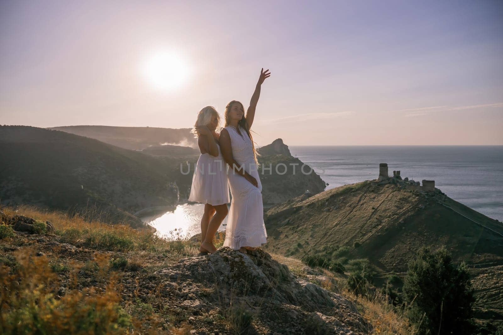 Two young girls are standing on a hillside, one of them wearing a white dress. The sun is shining brightly, creating a warm and inviting atmosphere. The girls seem to be enjoying their time together