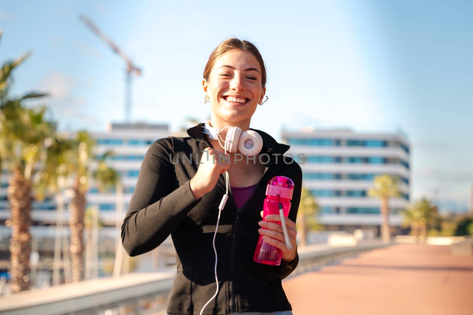 A smiling fitness woman runs with headphones and water bottle outdoors. Workout concept and healthy lifestyle.