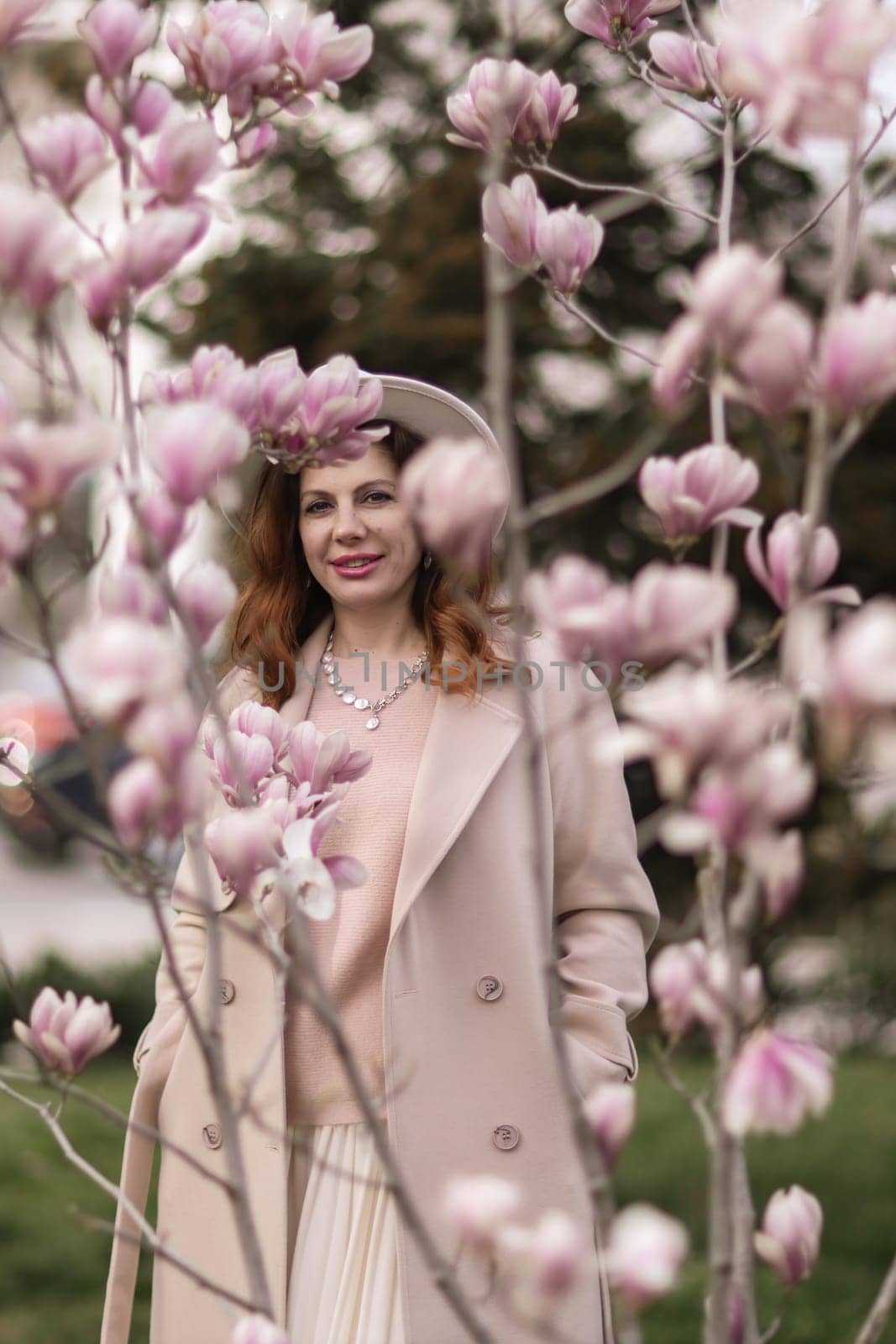 Woman magnolia flowers, surrounded by blossoming trees, hair down, white hat, wearing a light coat. Captured during spring, showcasing natural beauty and seasonal change