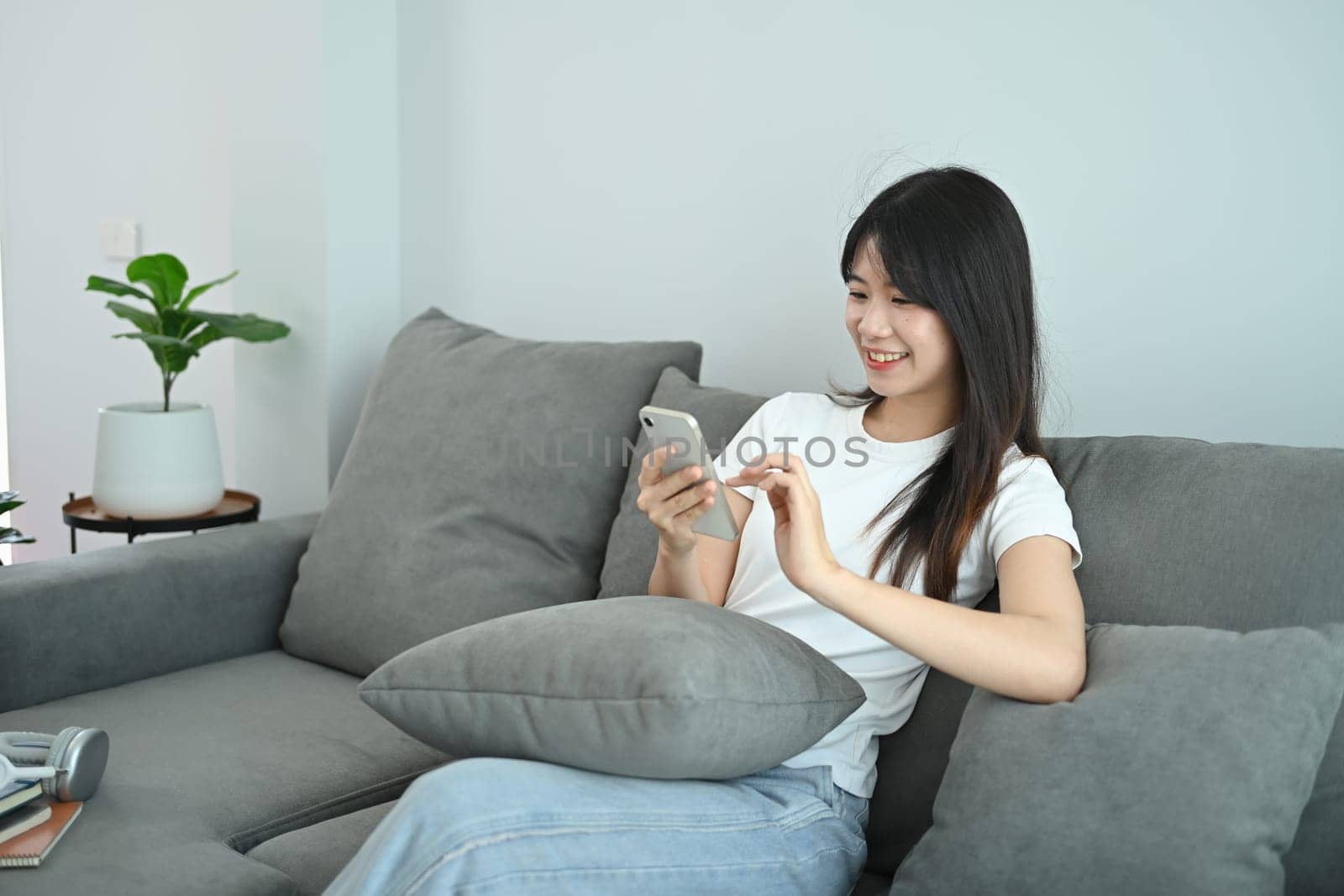 Smiling asian woman in casual clothes relaxing and using smartphone on couch.