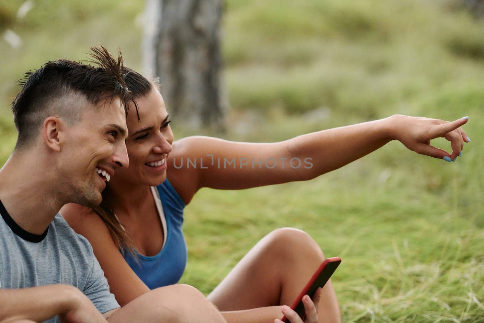 A romantic couple sits in the forest, browsing new running routes on their smartphone, embracing the beauty of nature as they plan their next outdoor adventure together