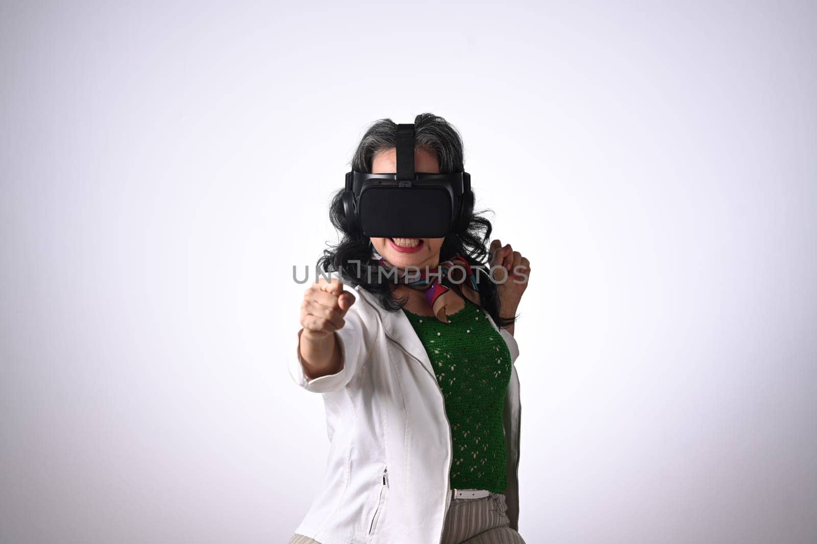 Joyful senior woman in boxing stance, playing action simulator game in VR headset.