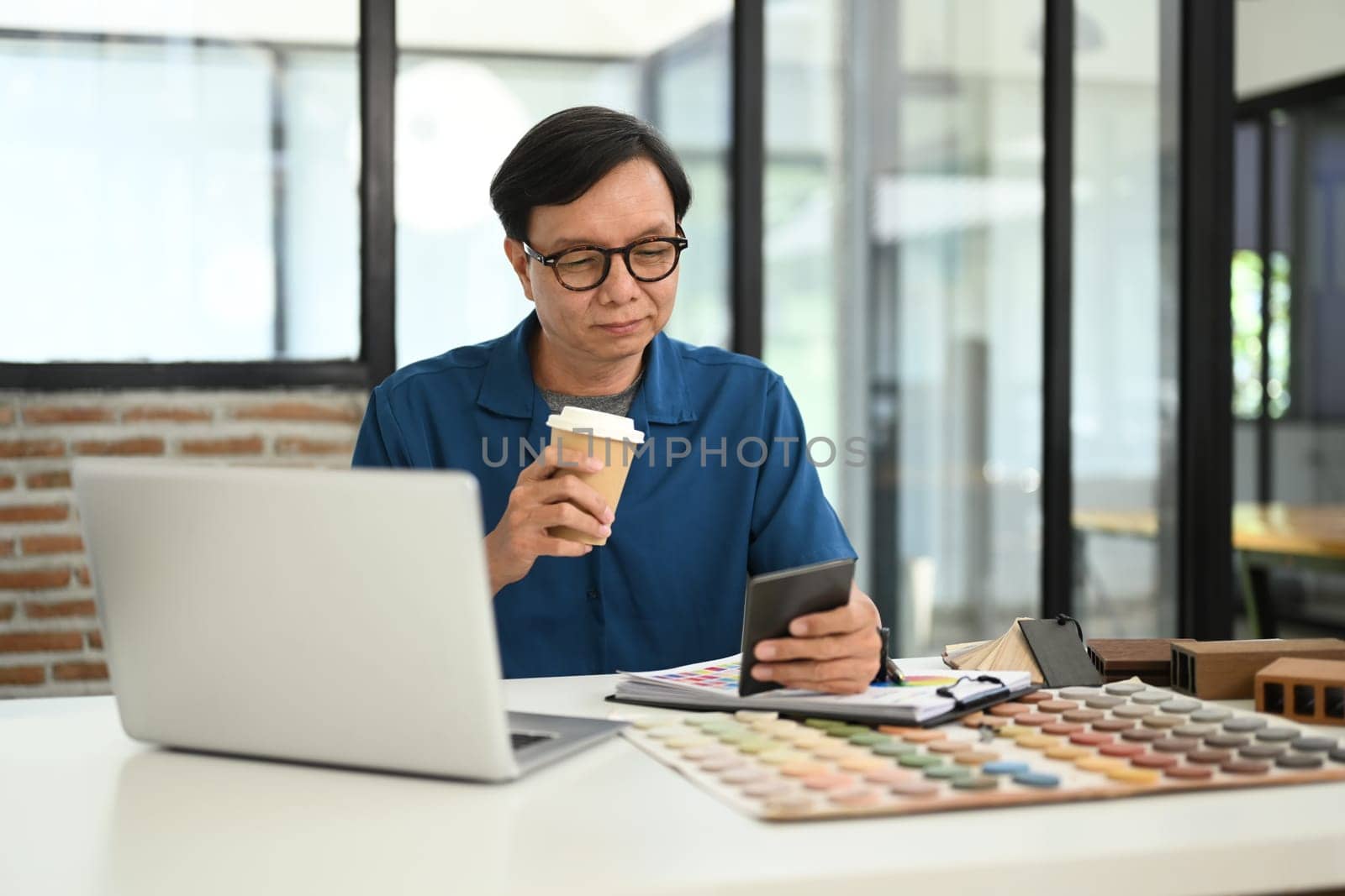 Portrait of senior male interior designer drinking coffee and using mobile phone at creative workplace.