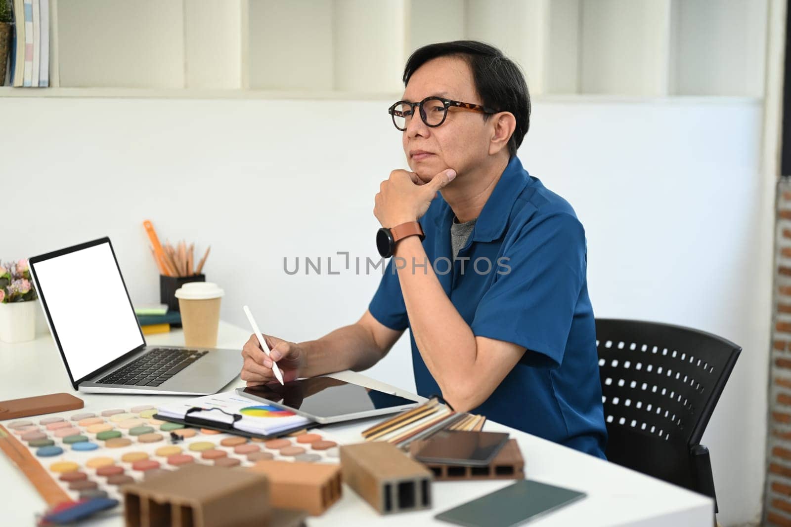 Thoughtful middle age interior designer sitting at workplace and looking away.