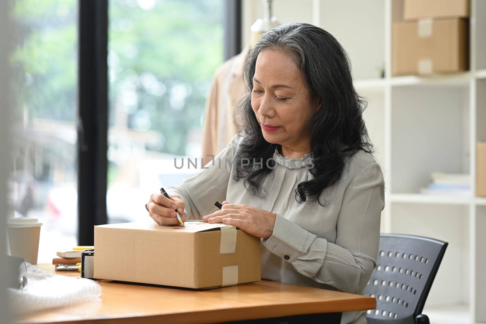 Middle aged woman entrepreneur writing address on cardboard for delivery to customers.