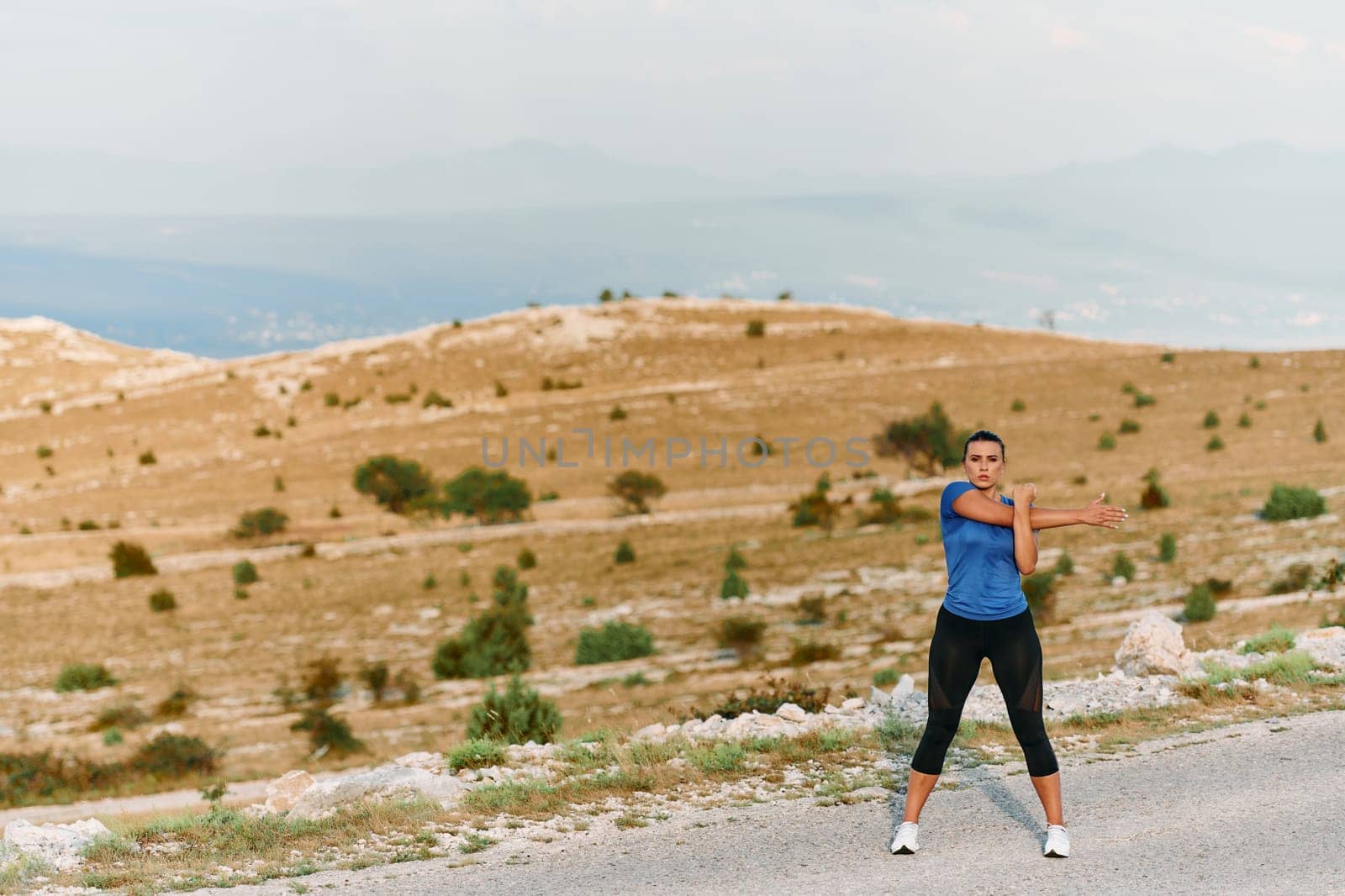 A determined female athlete stretches her muscles after a strenuous run through rugged mountain terrain, surrounded by breathtaking rocky landscapes.