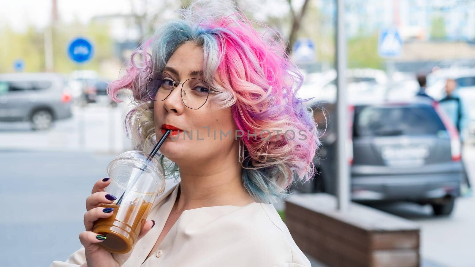 Close-up portrait of curly Caucasian woman with multi-colored hair wearing glasses. The hairstyle model is drinking a cold drink