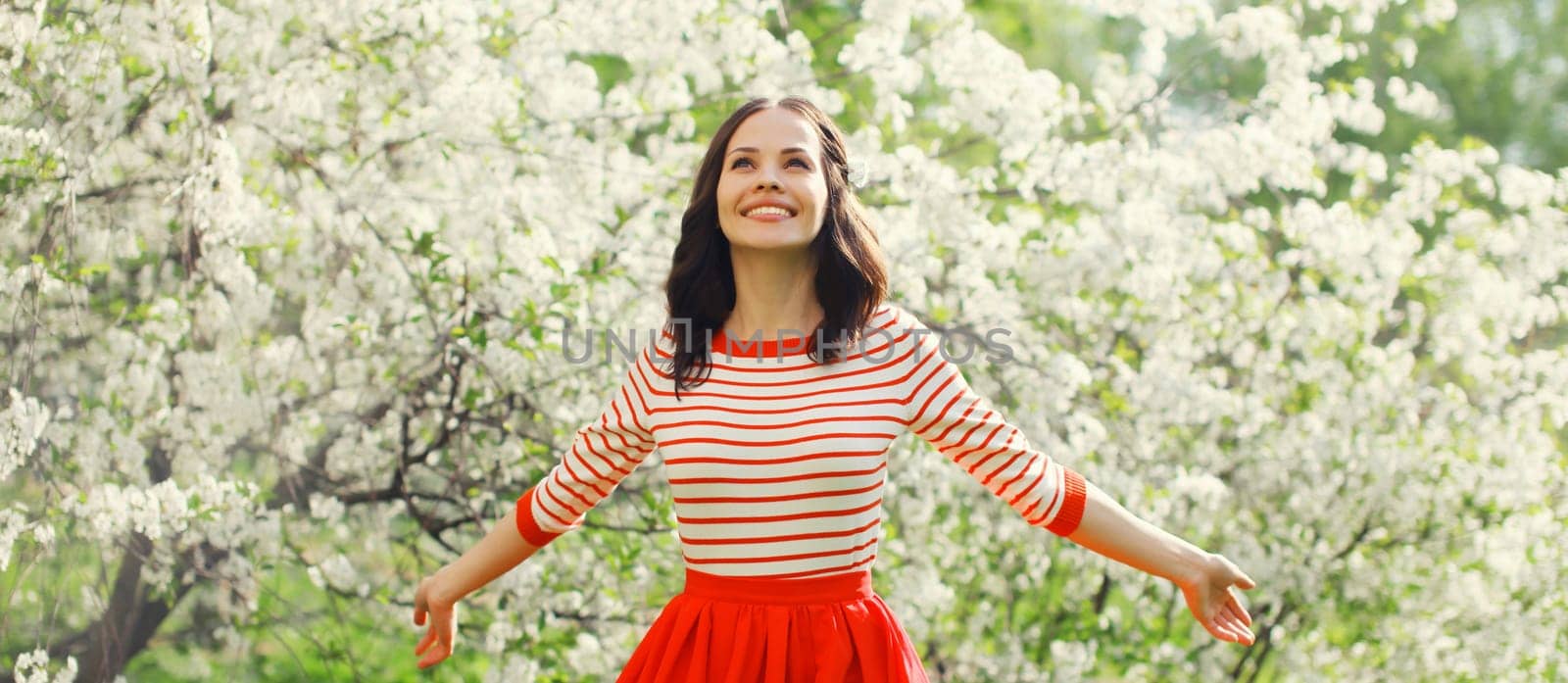 Lovely happy smiling young woman enjoying sunny day in spring blooming garden with white flowers on the trees in park