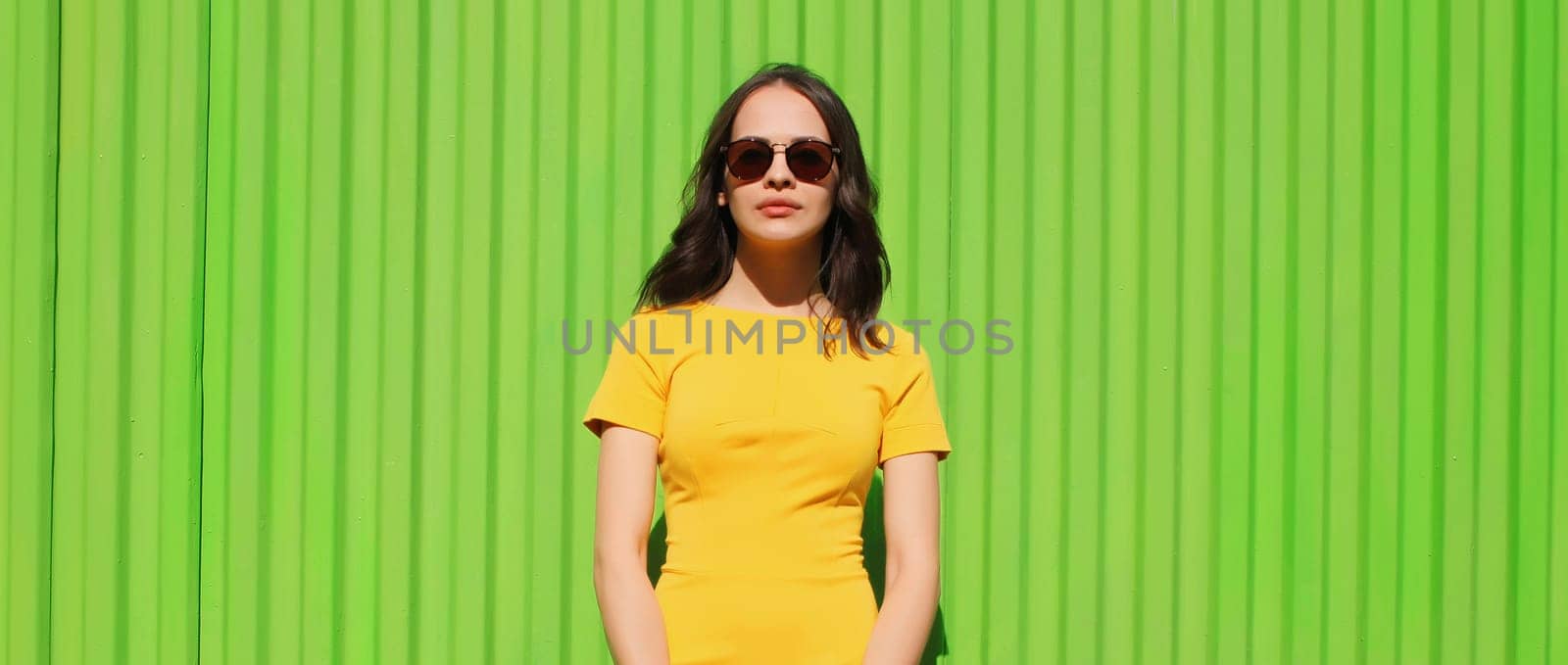 Portrait of beautiful young brunette woman model posing in yellow dress on city street on bright green background