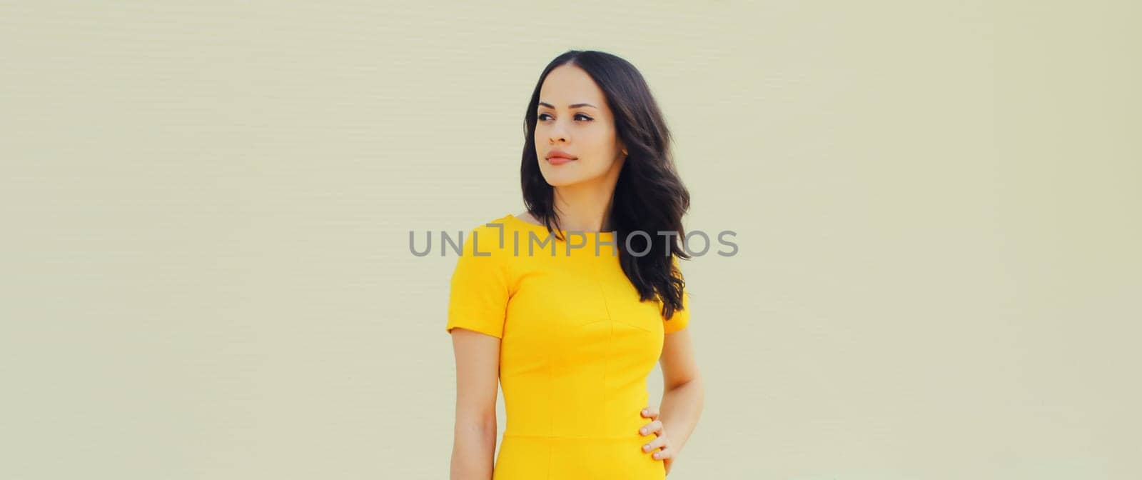 Portrait of beautiful young brunette woman model posing in yellow dress looking away on gray background