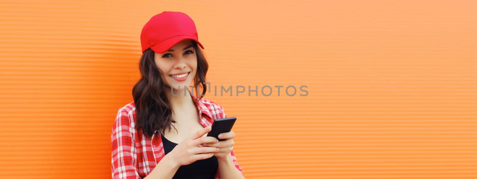 Portrait of happy smiling young woman with mobile phone in summer red baseball cap on colorful orange background, blank copy space for advertising text