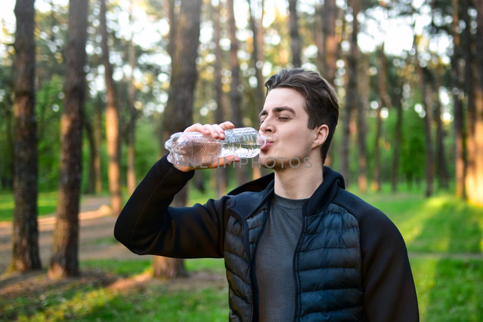 A handsome young man in a sports jacket drinking water from a bottle in nature.
