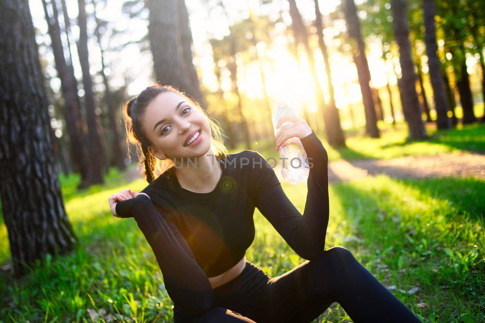 A smiling young woman in a sporty black suit holds a water bottle in the woods