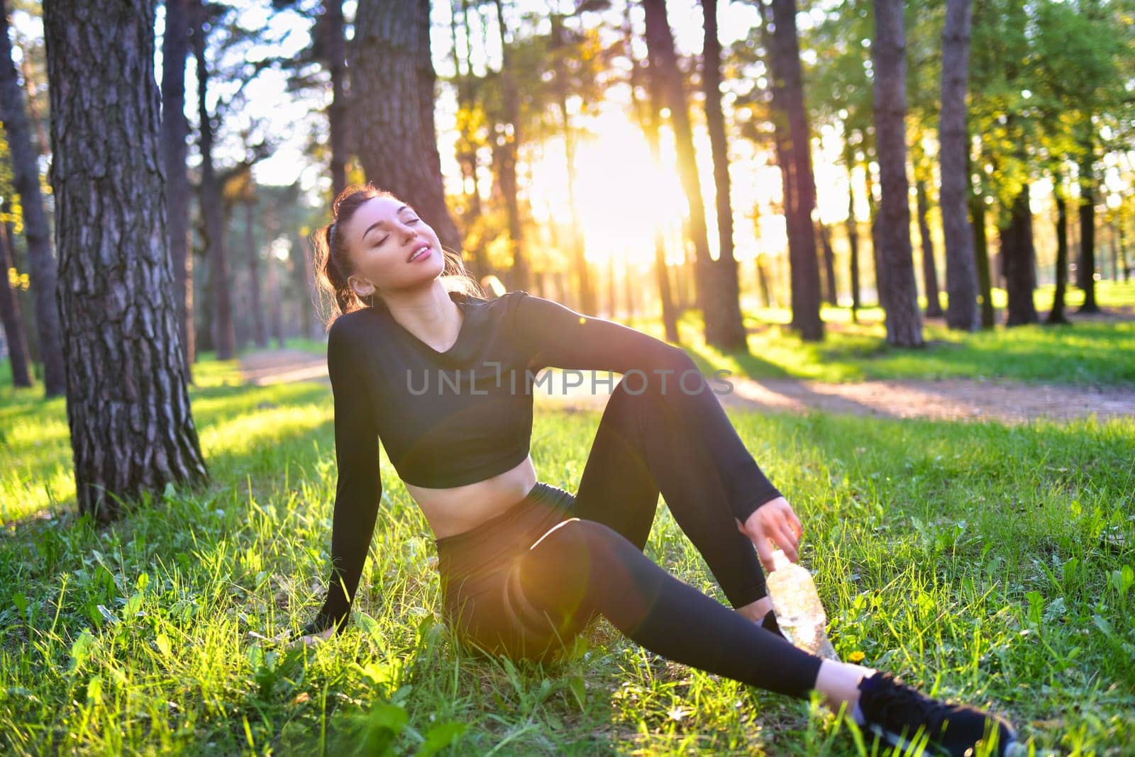 A smiling young woman in a sporty black suit holds a water bottle in the woods.