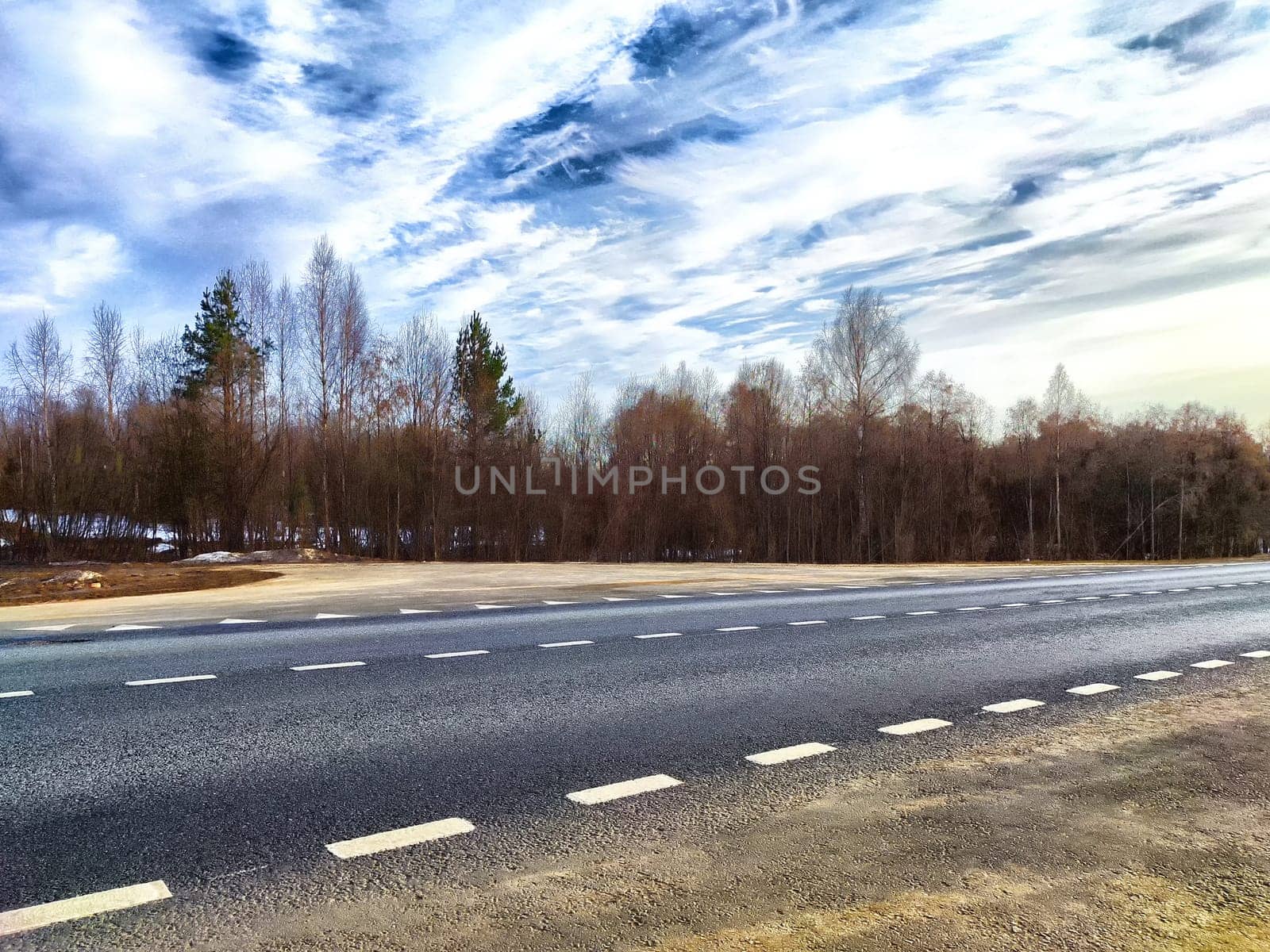 A lonely country road stretches alongside barren trees under a clear, partly cloudy sky on peaceful winter afternoon