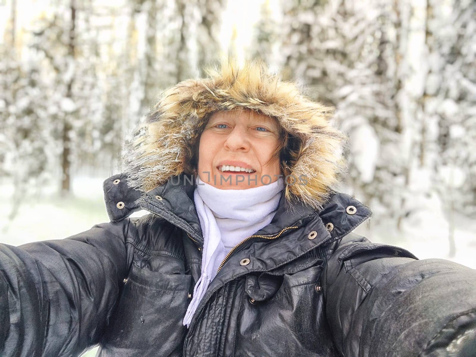 A cheerful middle aged woman in a winter coat with fur, scarf taking selfie on nature outdoors and cold, snowy background with blue sky and white clouds