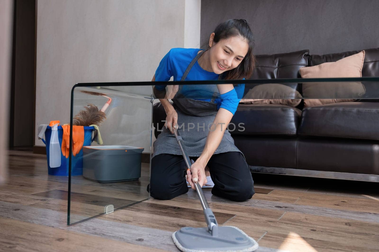 Smiling housekeeper mopping floor in living room with cleaning supplies. Concept of domestic chores and hygiene by wichayada