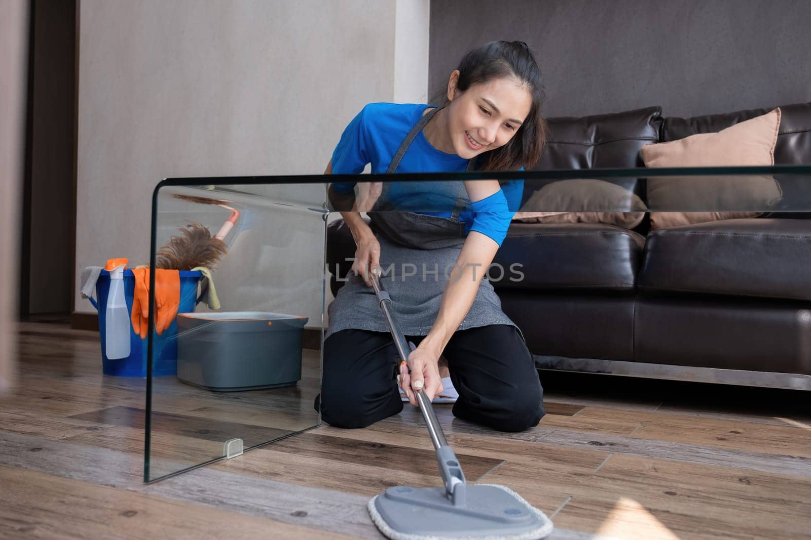 Smiling housekeeper mopping floor in living room with cleaning supplies. Concept of domestic chores and hygiene.