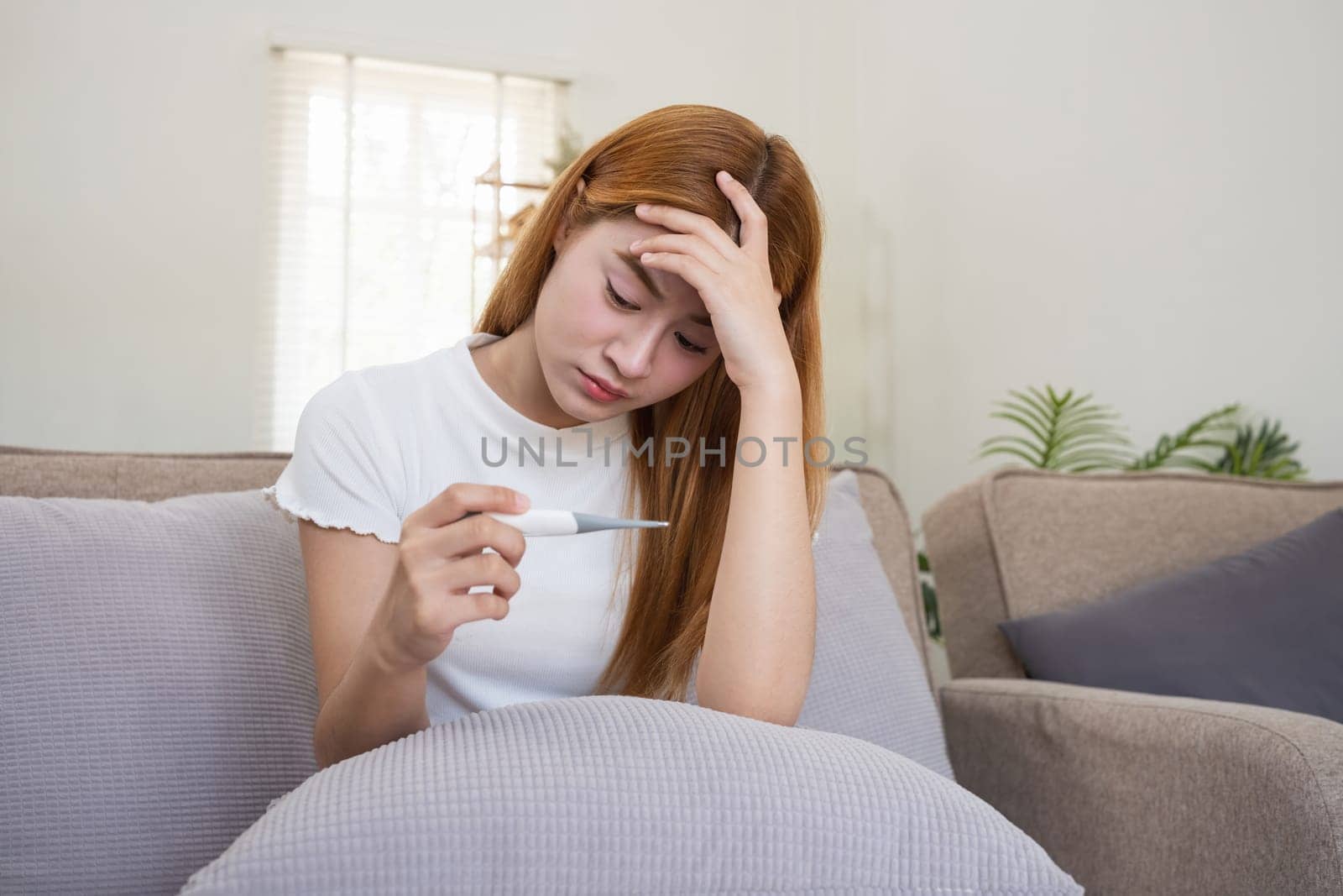 Young Asian woman with a fever, checking temperature at home. Concept of illness, fever, health issues, and self-care.