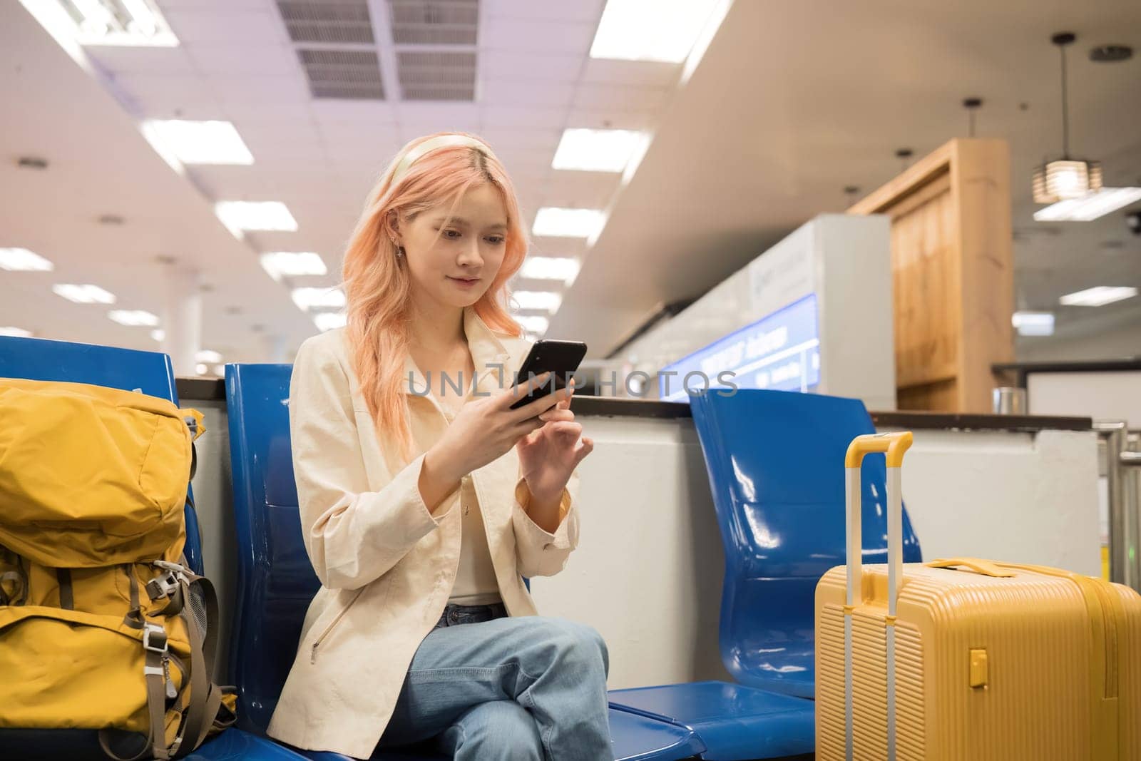 Asian woman using smartphone in airport with luggage. Concept of travel, technology, and waiting.