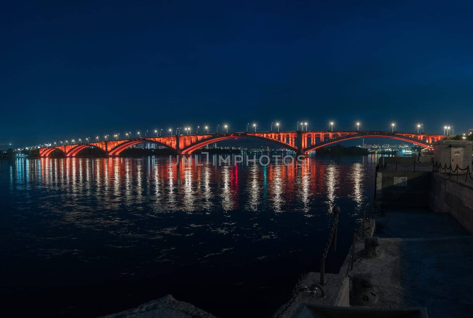 A bridge crossing over a body of water during the nighttime, set against a cityscape with the sky and horizon visible in the background