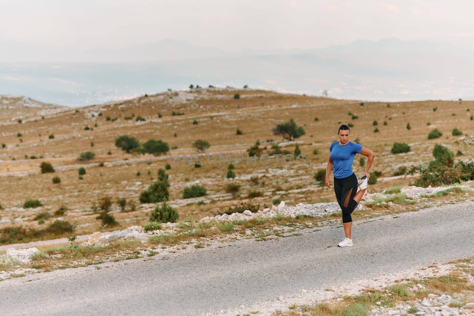 A determined female athlete stretches her muscles after a strenuous run through rugged mountain terrain, surrounded by breathtaking rocky landscapes.