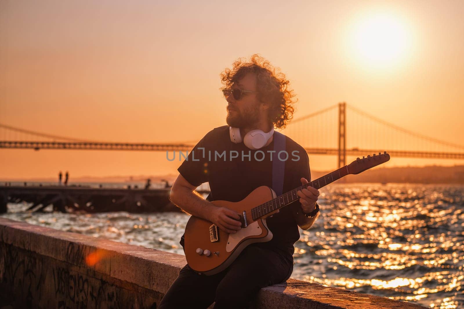 Hipster street rock musician in black playing electric guitar in the street on sunset on embankment with 25th of April bridge in background. Lisbon, Portugal