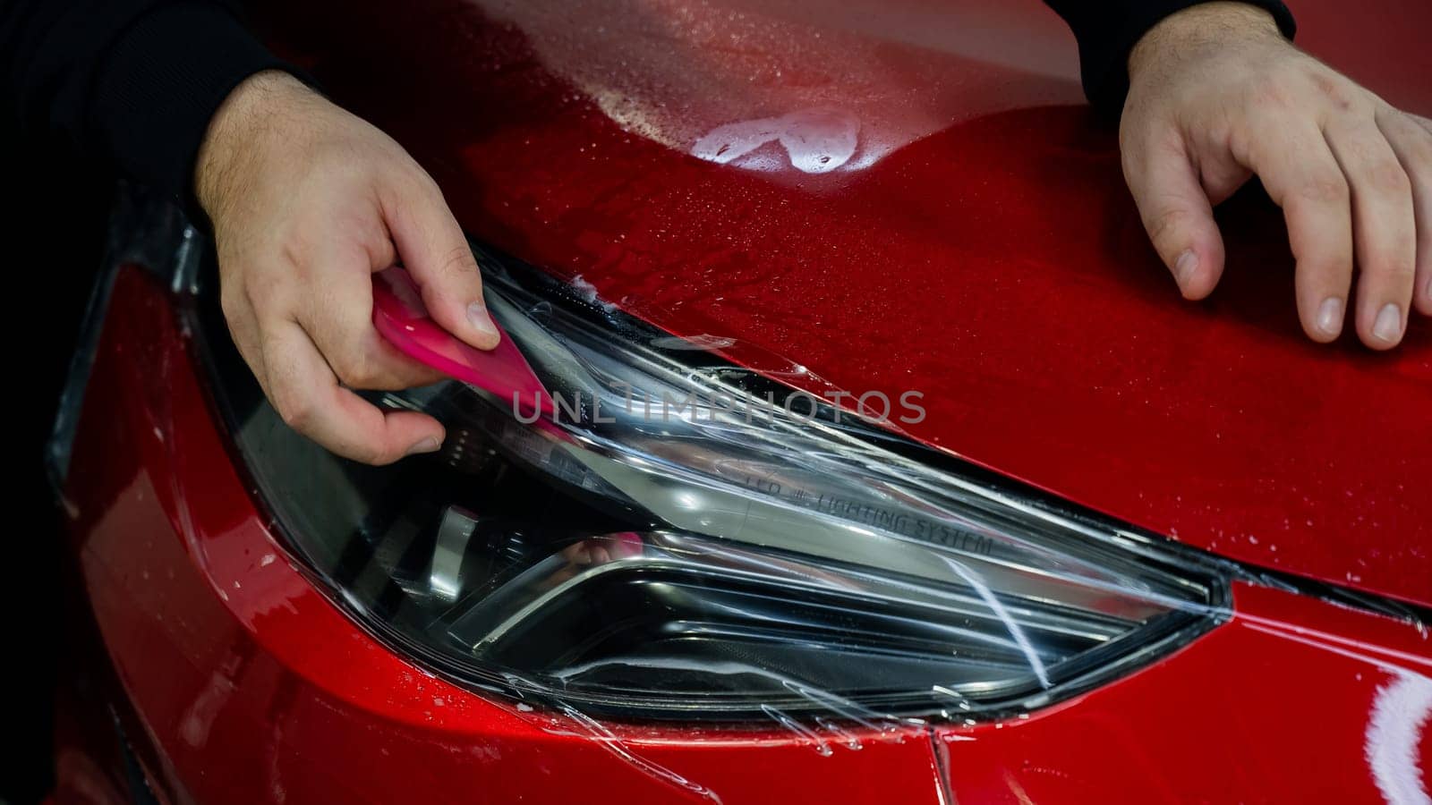 The master applies vinyl film to the headlight of a red car. Closeup view on worker detailer hand smoothing with a scraper protective film