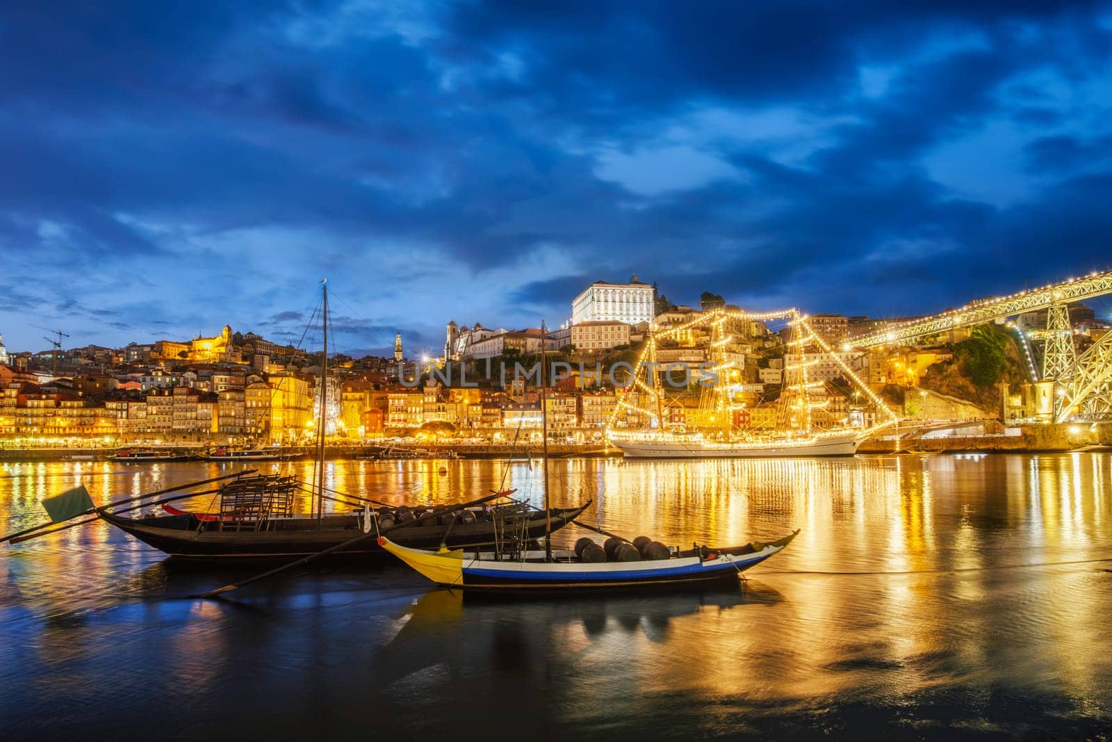 View of Porto city and Douro river with traditional boats with port wine barrels and sailing ship from famous tourist viewpoint Marginal de Gaia riverfront in night. Porto, Vila Nova de Gaia, Portugal