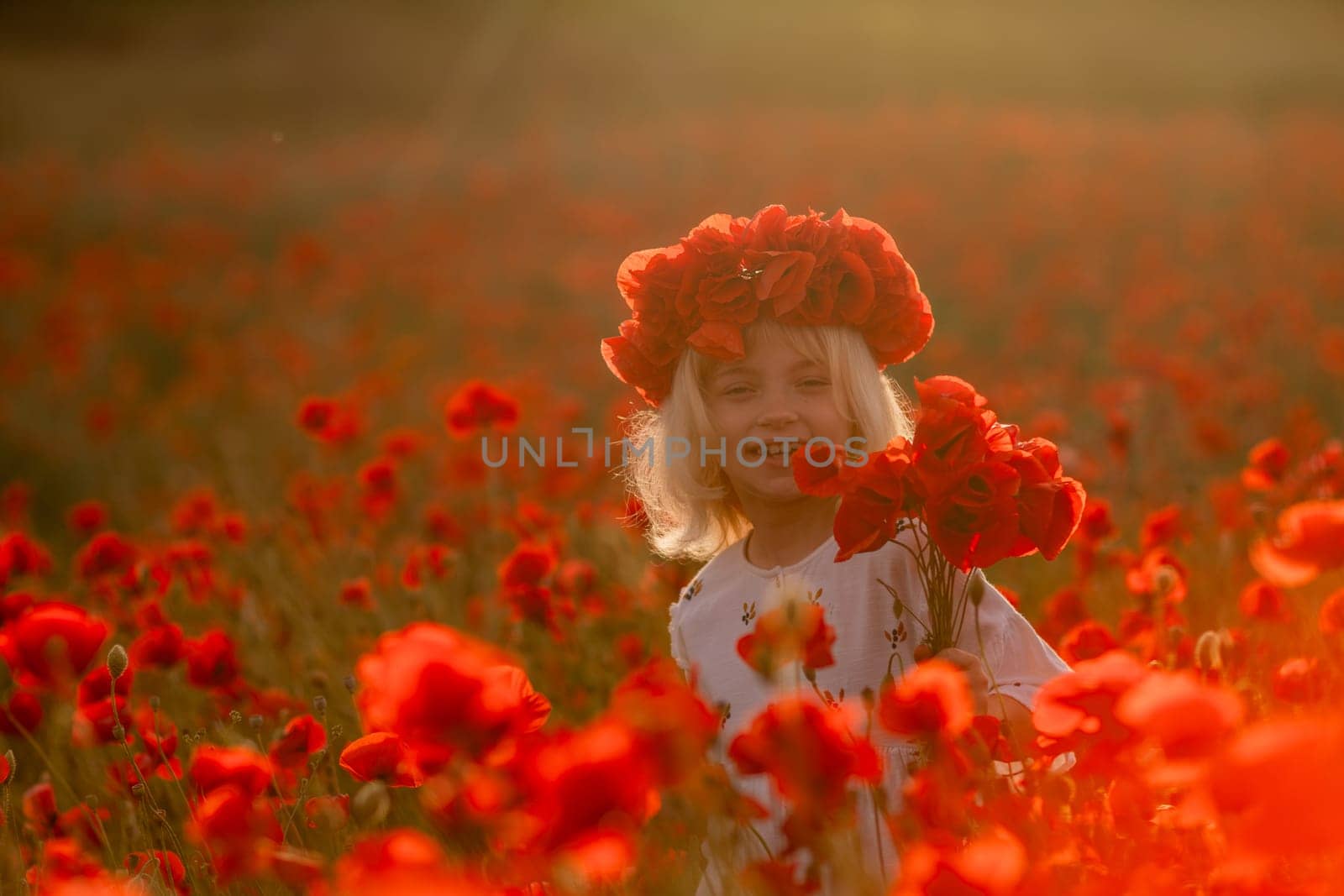 A young girl is standing in a field of red poppies. She is wearing a red headband and holding a bouquet of flowers. The scene is bright and cheerful