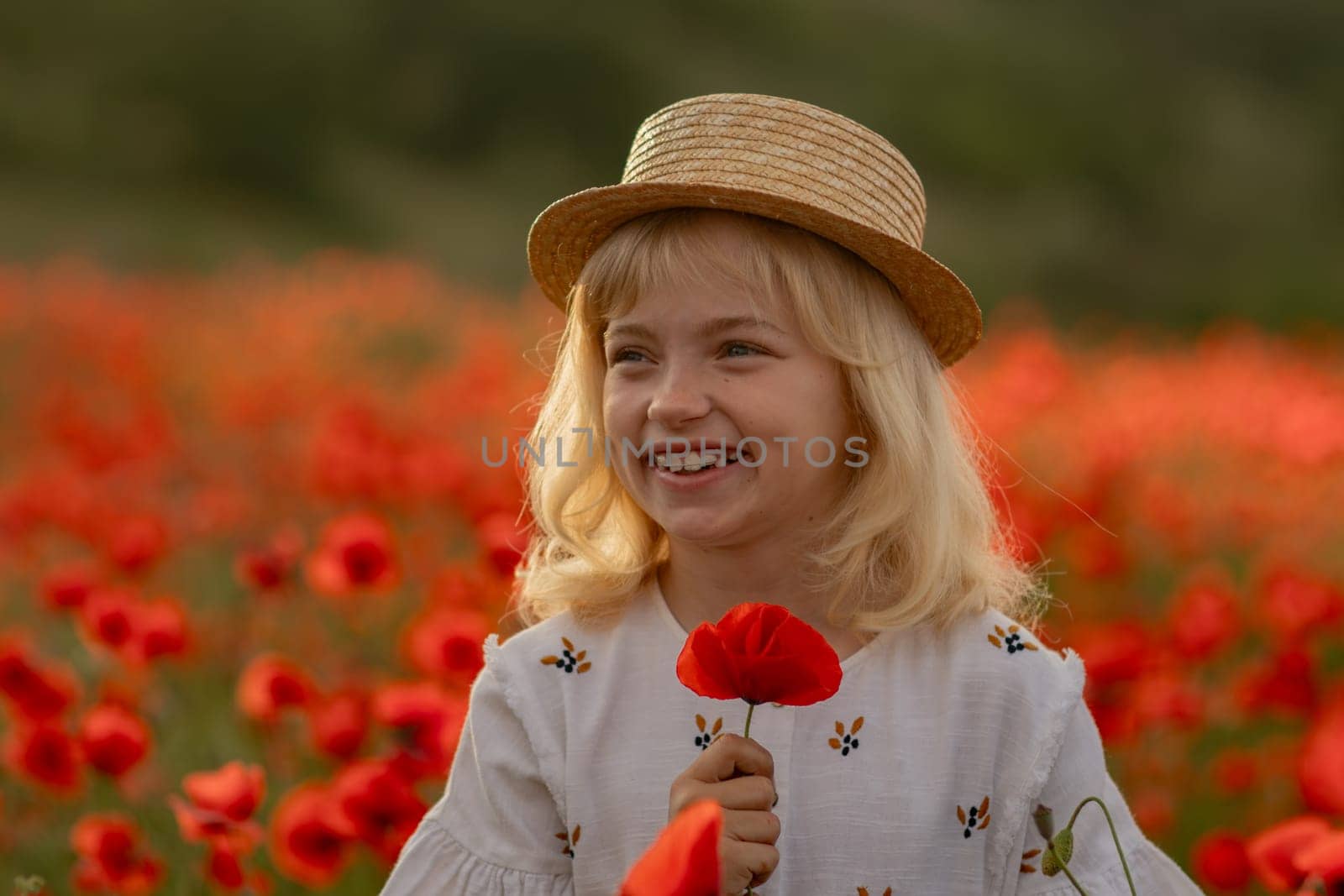 A young girl is standing in a field of red poppies, holding a red flower. She is smiling and she is enjoying the beautiful scenery