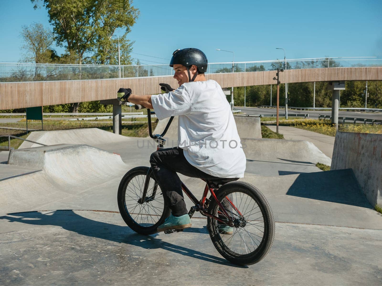 Smiling young bmx rider sitting on bmx bike in skatepark. Side view full body portrait of happy smiling bmx rider in protection helmet, gloves and leg protection. Young BMX bicycle rider having fun and posing