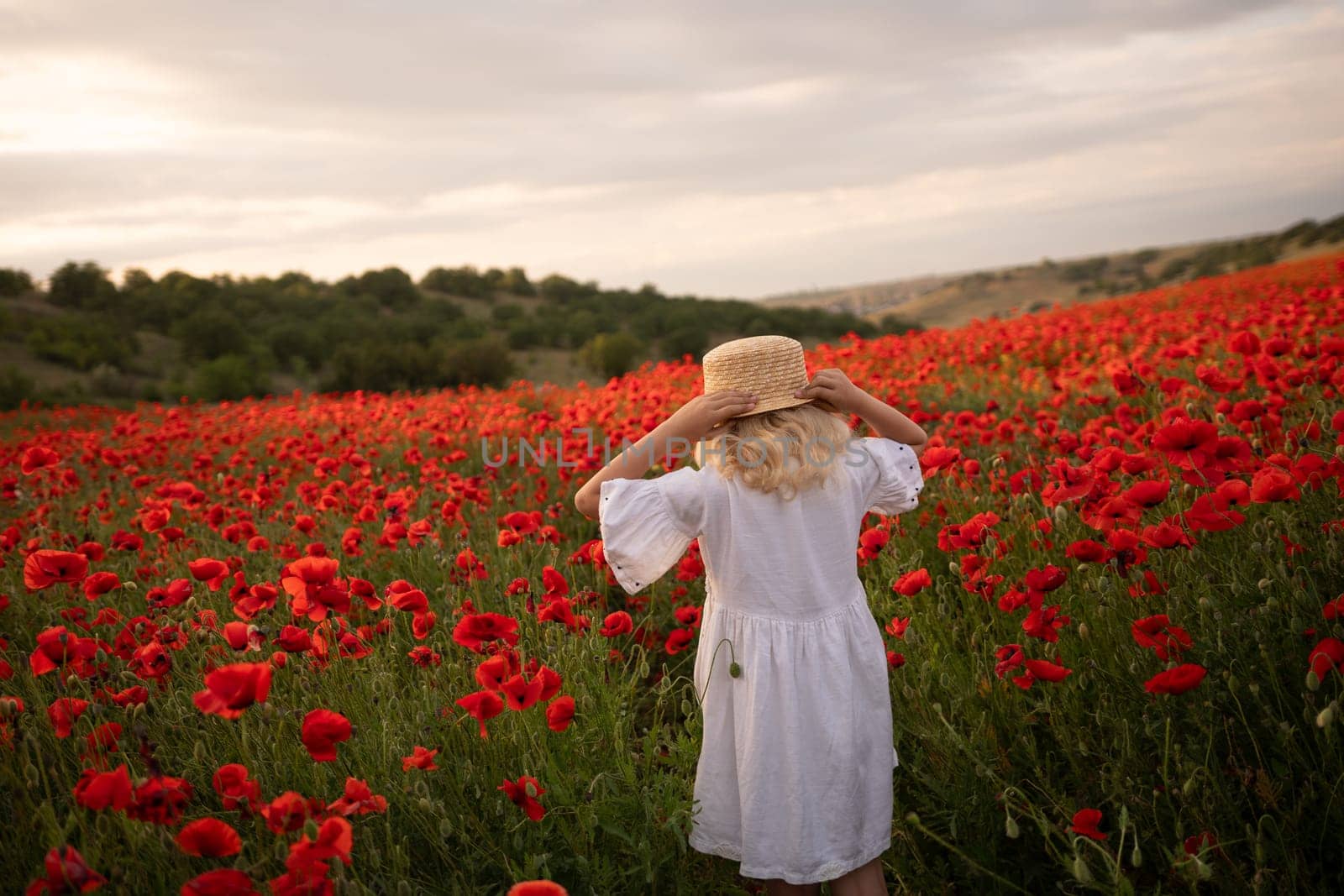A young girl wearing a straw hat stands in a field of red poppies. The scene is serene and peaceful, with the girl looking out over the vast expanse of flowers