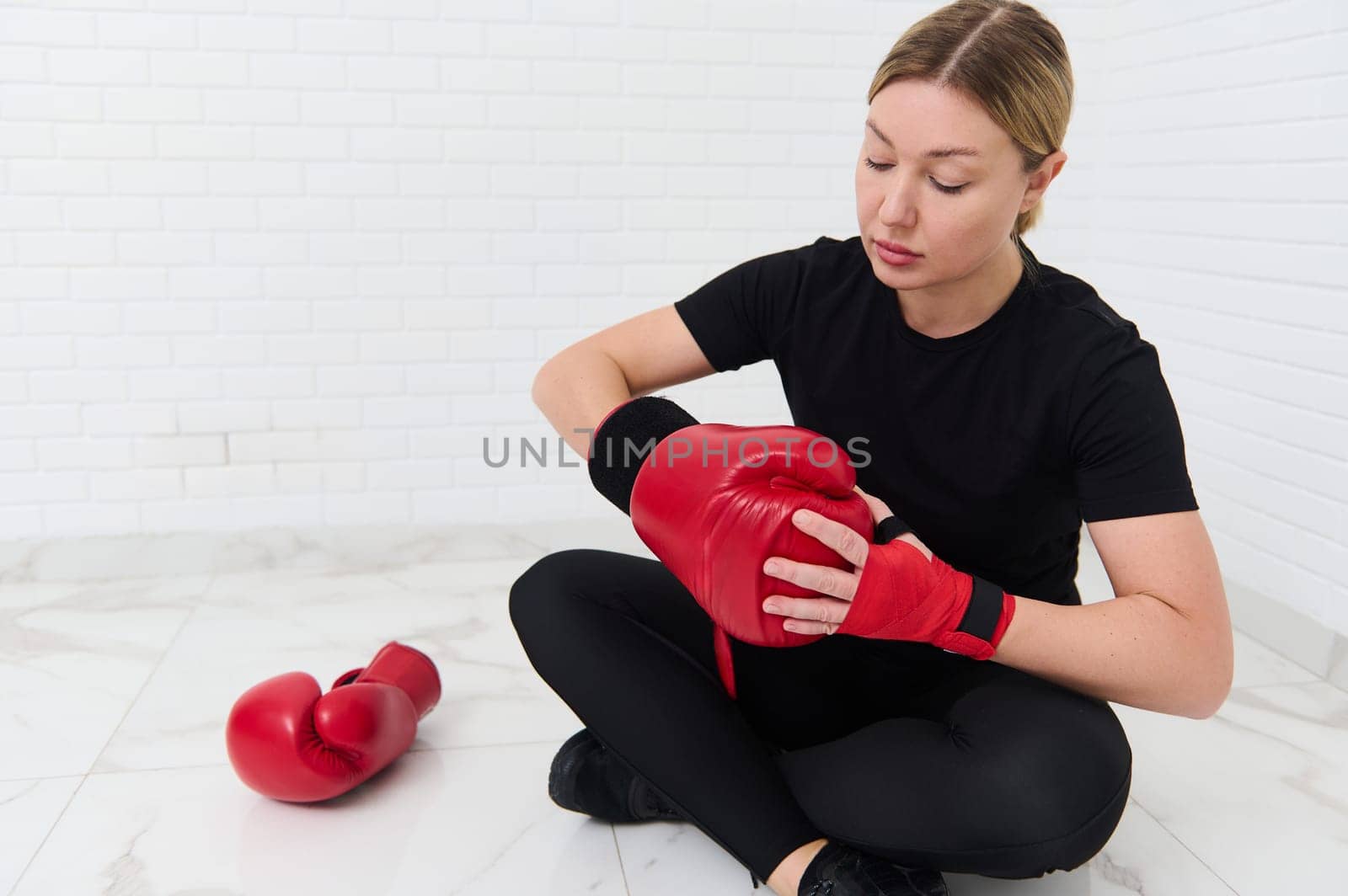 Portrait of European woman boxer putting on red boxing gloves, sitting against white wall background. The concept of athletic discipline, sport, combat, martial art.