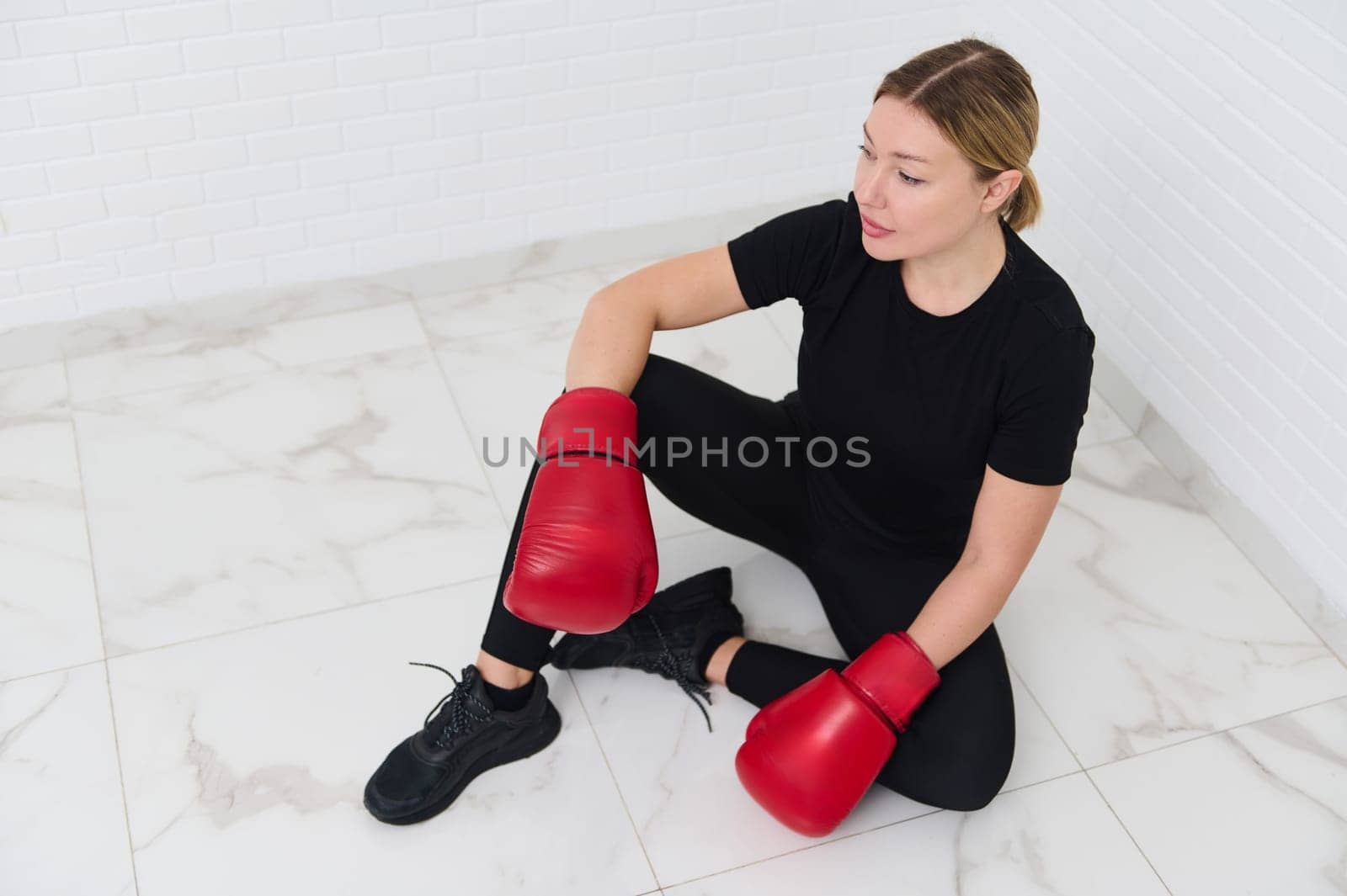 View from above of a blonde female boxer fighter wearing red boxing gloves, getting ready for combat, sitting over white wall background. Martial art combat. People and sport