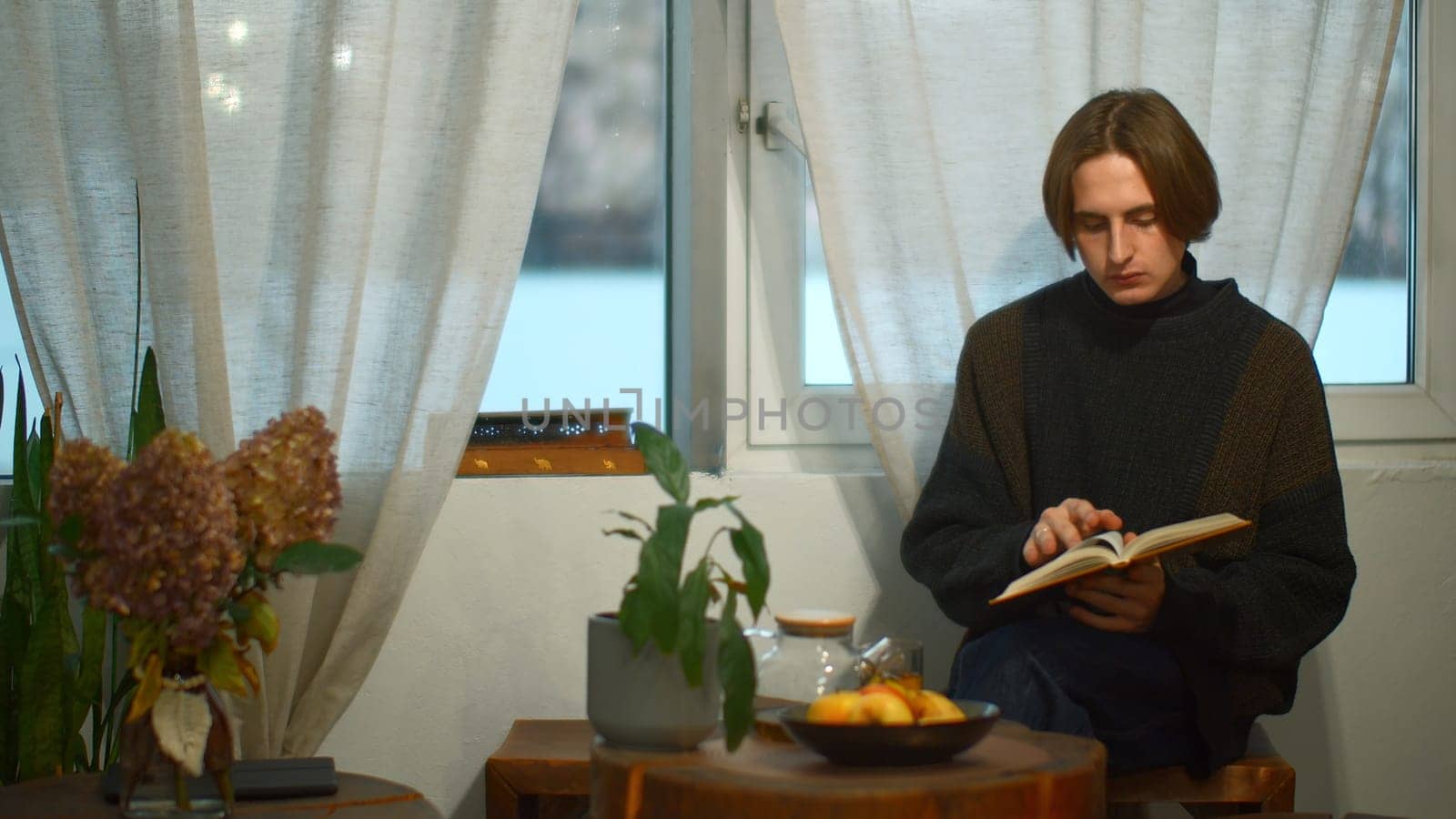 Young handsome man is reading book with tea in cafe. Media. Student is sitting alone and reading book in cozy cafe. Young man is reading book and drinking tea in student cafe.