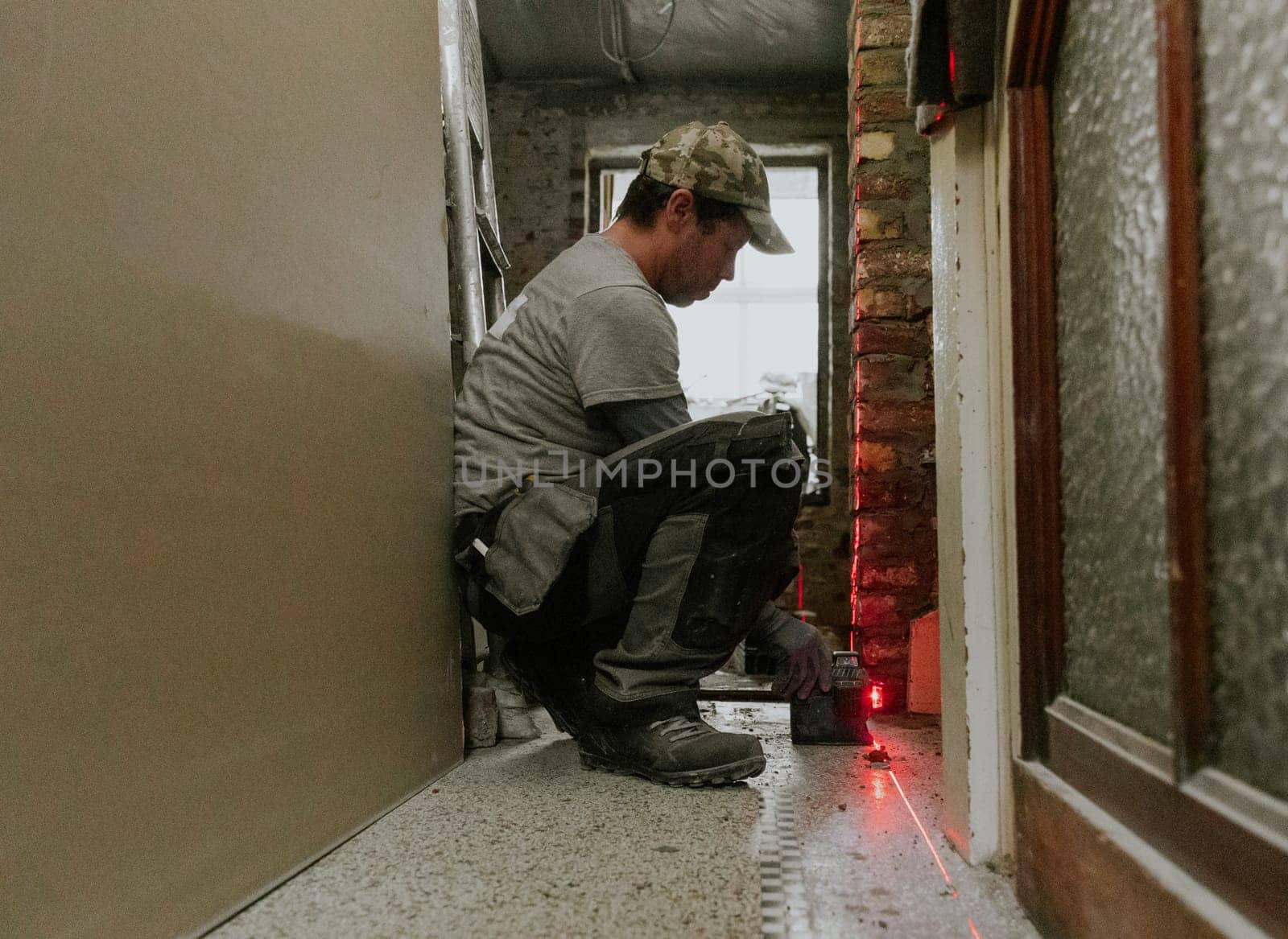 One young Caucasian man in casual clothes squats, looks down and measures fresh brickwork in the doorway of a long corridor in an old house using a laser level, close-up view from below.