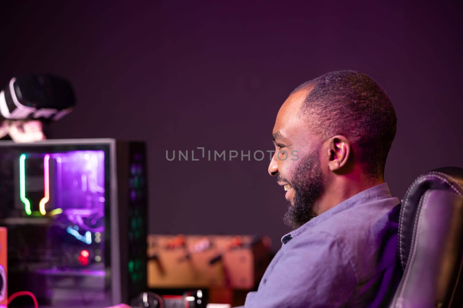 Gamer laughing at home, having fun playing videogames on computer. Joyous african american man contending in gaming tournament using powerful neon lit desktop PC in apartment