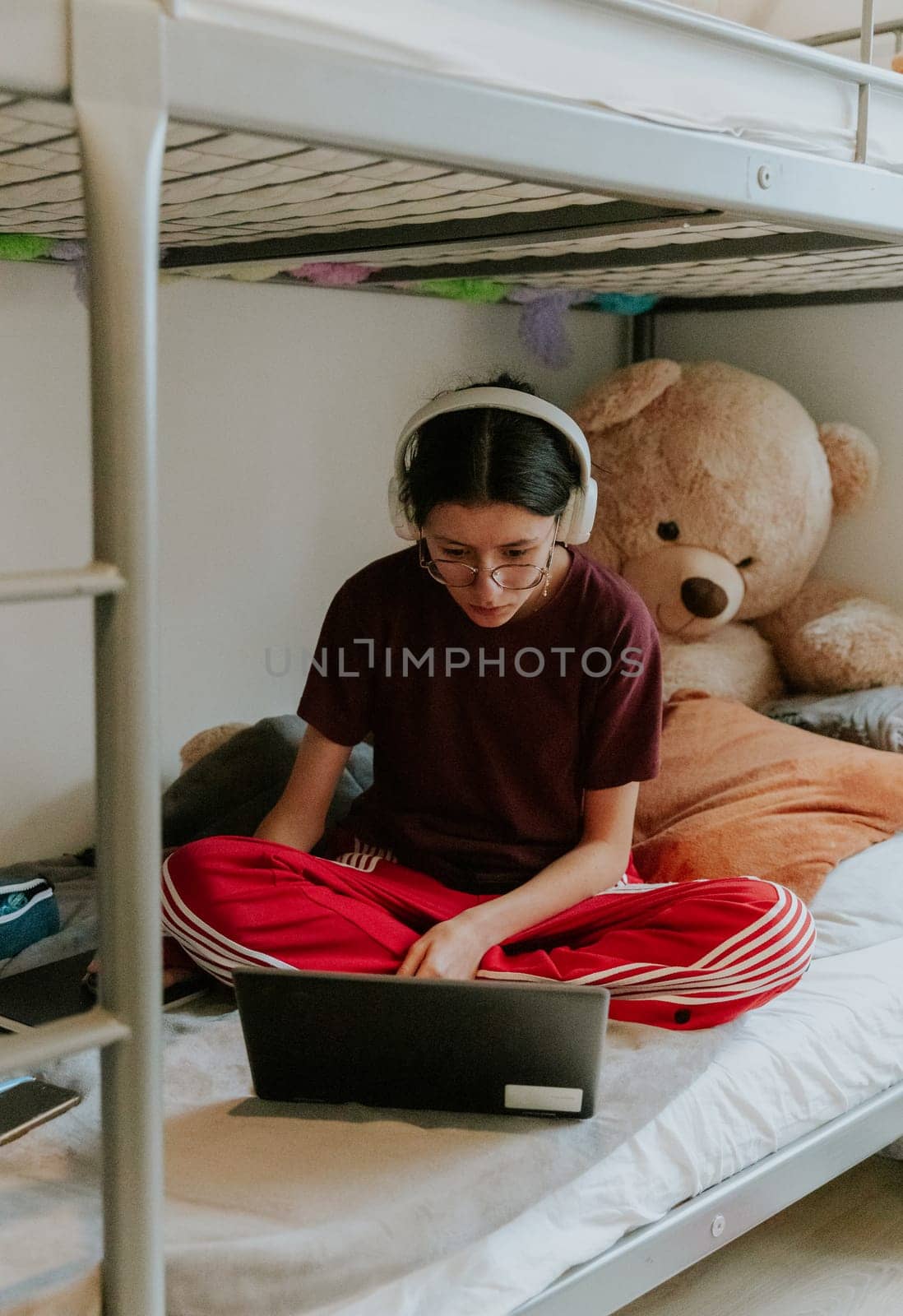 One young beautiful Caucasian teenage girl with white headphones and sportswear sits on a bunk bed with a large toy bear and teaches homework at the computer, side view close-up.