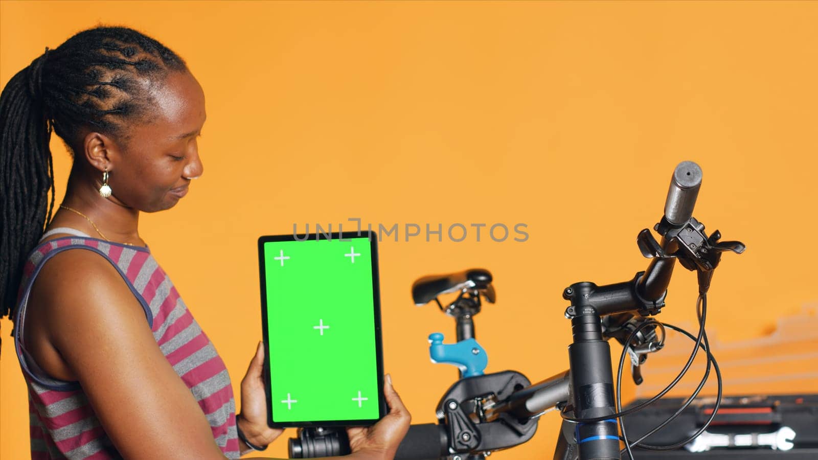 Woman holds green screen tablet, using it to advertise bike repair shop, empty placeholder for company logo, studio background. Mechanic promoting bicycle fixing service with mockup device, camera A