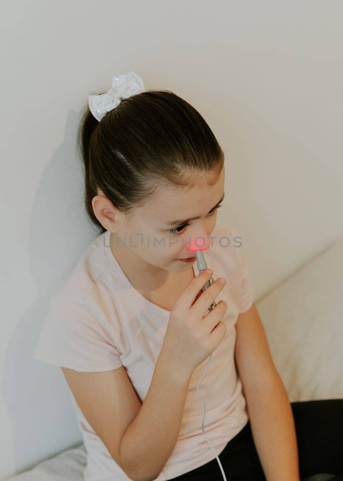 One beautiful Caucasian brunette girl with a serious emotion, collected hair and in a pink T-shirt treats the right nasal passage with a device with infrared light, sitting on the bed, cross-legged, leaning on a white wall, close-up view from above.