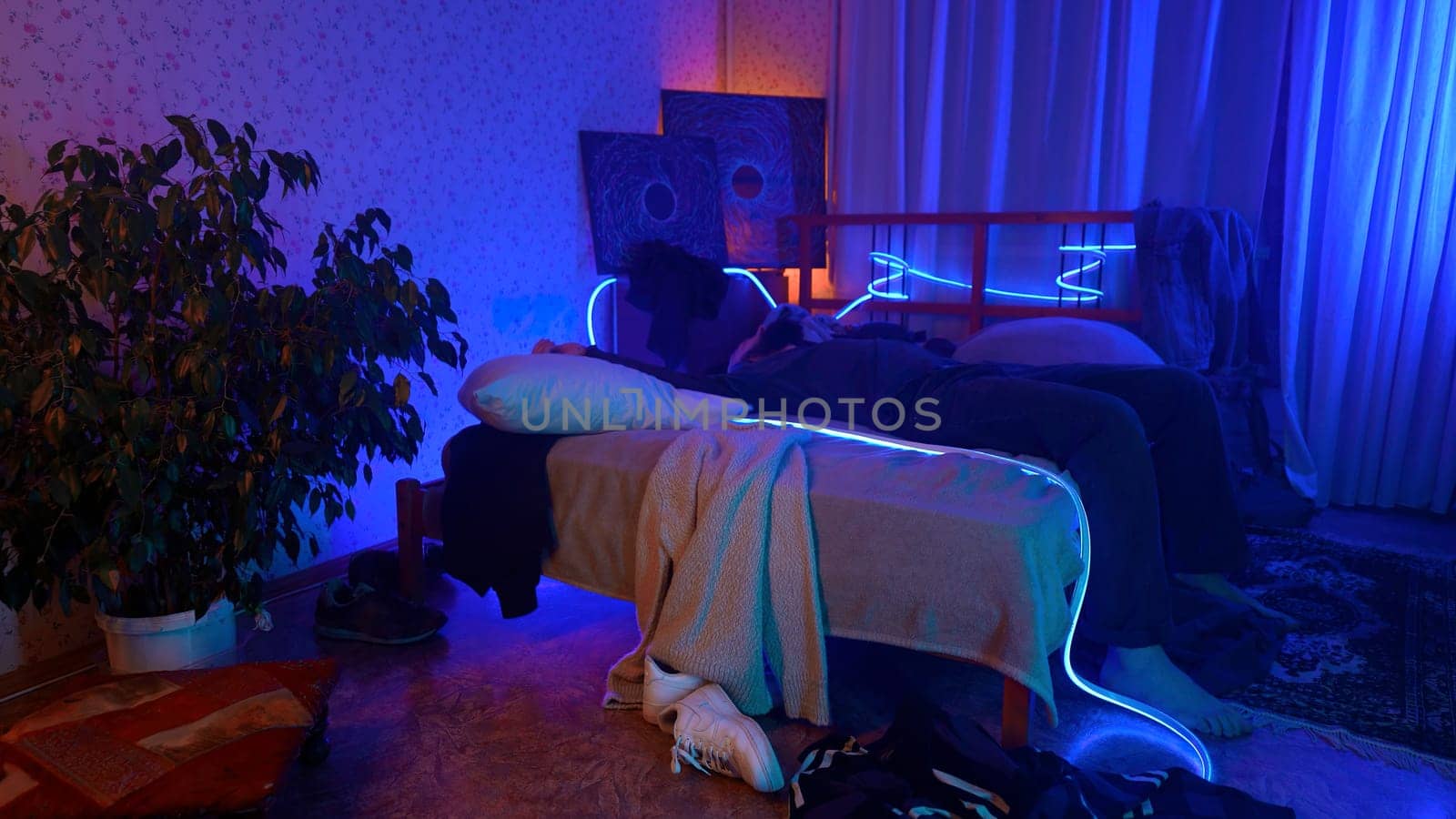 Worried, tired man sitting on bed at night and listening to music. Media. Young man can't sleep at night in a room with neon lighting