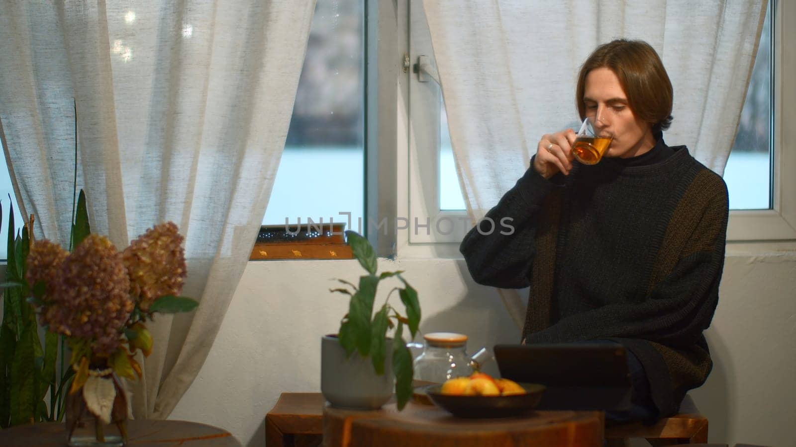 Young man drinks tea and looks at tablet in cafe. Media. Handsome young man is drinking tea alone and looking at tablet. Student is relaxing with tablet in cozy cafe.