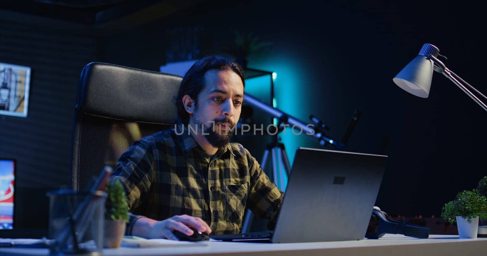 Freelancer in neon lit home office sitting at desk and typing on laptop keyboard. Smiling remote employee using electronic device to do customers relations job online, answering inquiries, camera B