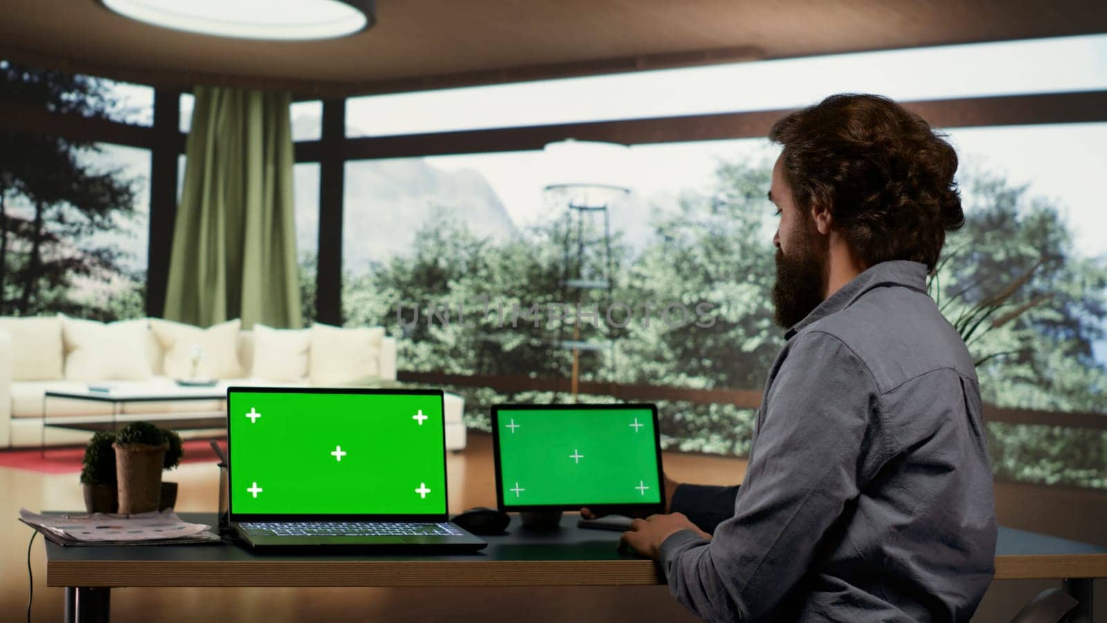 Businessperson using greenscreen displays on devices, sitting at his desk in luxurious villa in a forest. Rich entrepreneur looking at screens showing isolated copyspace, works on laptop and tablet.