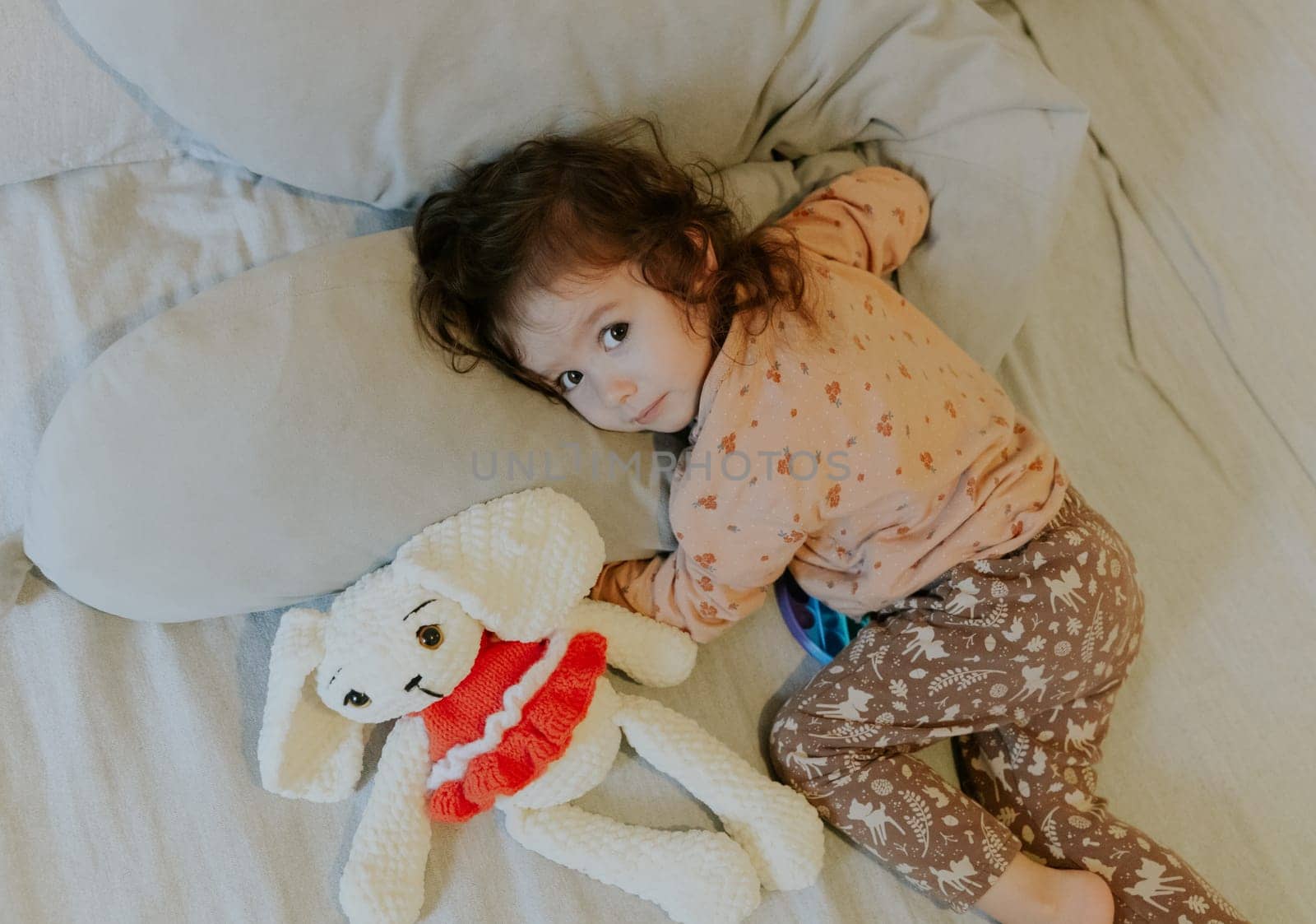 One beautiful little Caucasian girl lies on her stomach on the bed with a soft toy in the early morning in the room and looks at the camera, close-up top view.