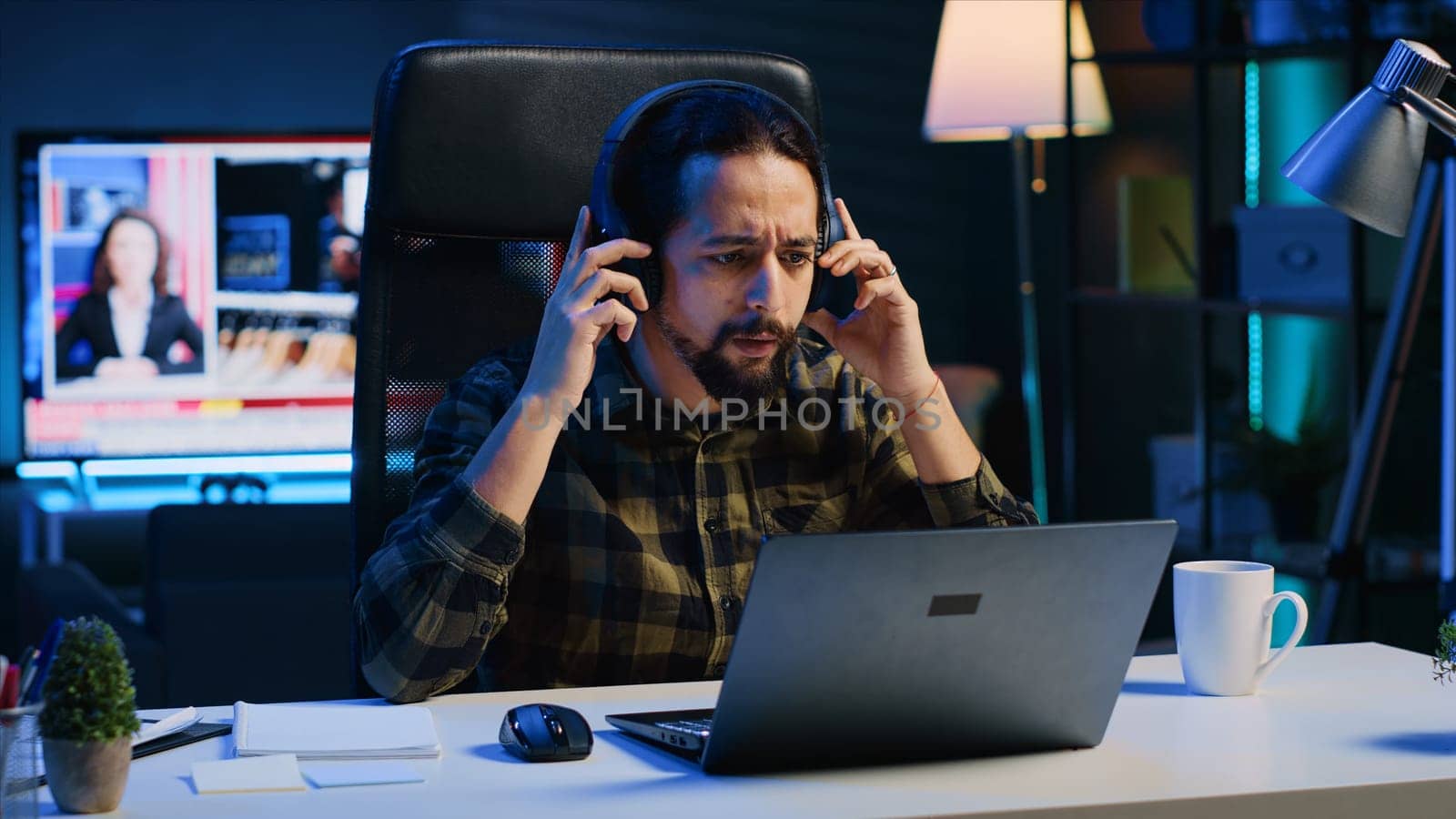 Freelancer enjoying music while researching data for company project, doing tasks in personal home office overnight. Remote worker sitting at desk in front of laptop and using headphones, camera A